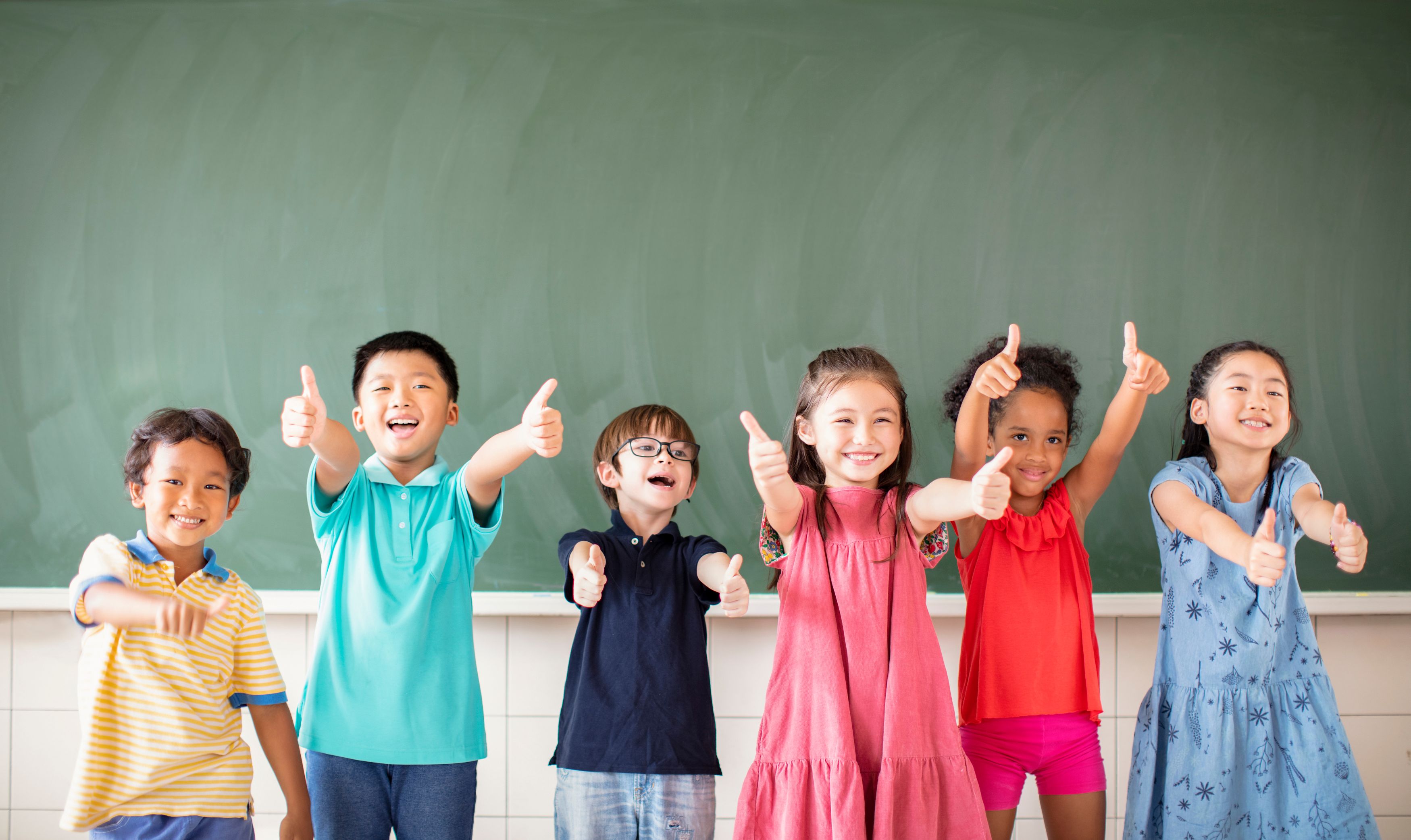Children in a classroom smile and give a thumbs-up while sitting at their desks in a bright, colorful learning environment.