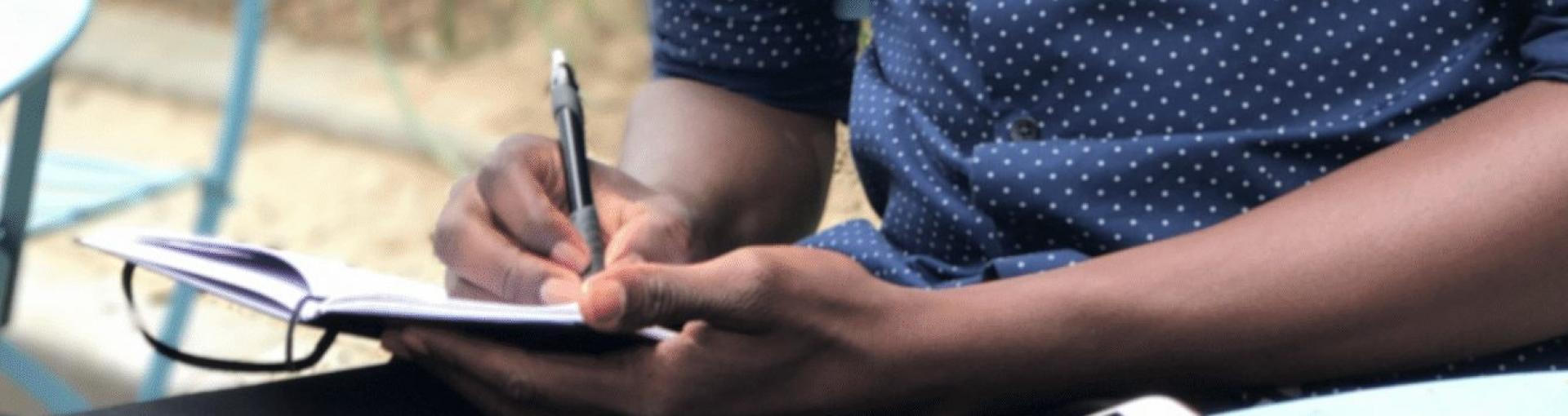 A close-up of a person writing in a notebook while sitting outdoors. They are holding a pen in their right hand and the notebook in their left, with glasses resting on the open pages. The person is wearing a blue shirt with small white polka dots.