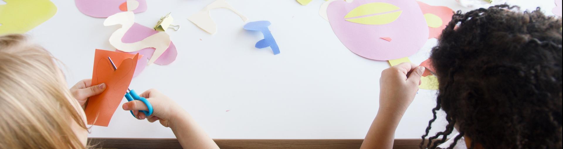A child with curly hair sits at a table working on a colorful paper craft project. They are arranging cut-out shapes, including a pink face with yellow lips, circles, and other abstract shapes, on a white surface. A couple of small containers and a binder clip are also on the table.