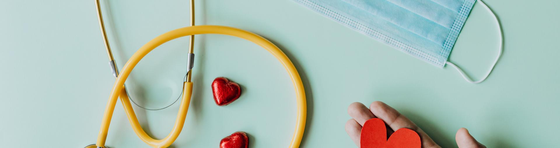 A yellow stethoscope, two red heart-shaped objects, a surgical mask, and a hand holding a red paper heart on a light green background.